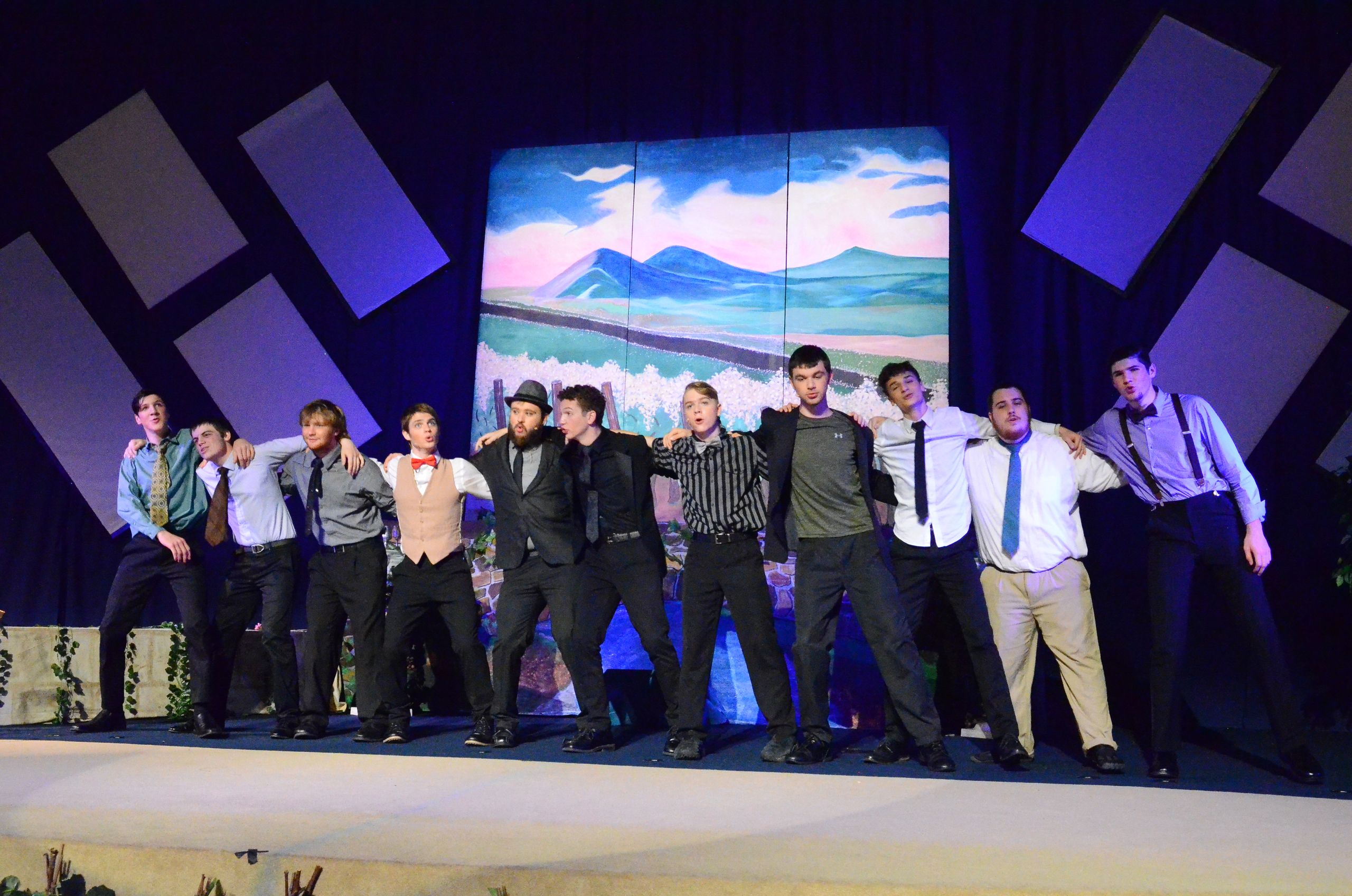 A cast of ten performers stands in a line across a stage wearing formal attire, with their arms around each other's shoulders, positioned in front of a large backdrop displaying a scenic landscape with mountains and water.