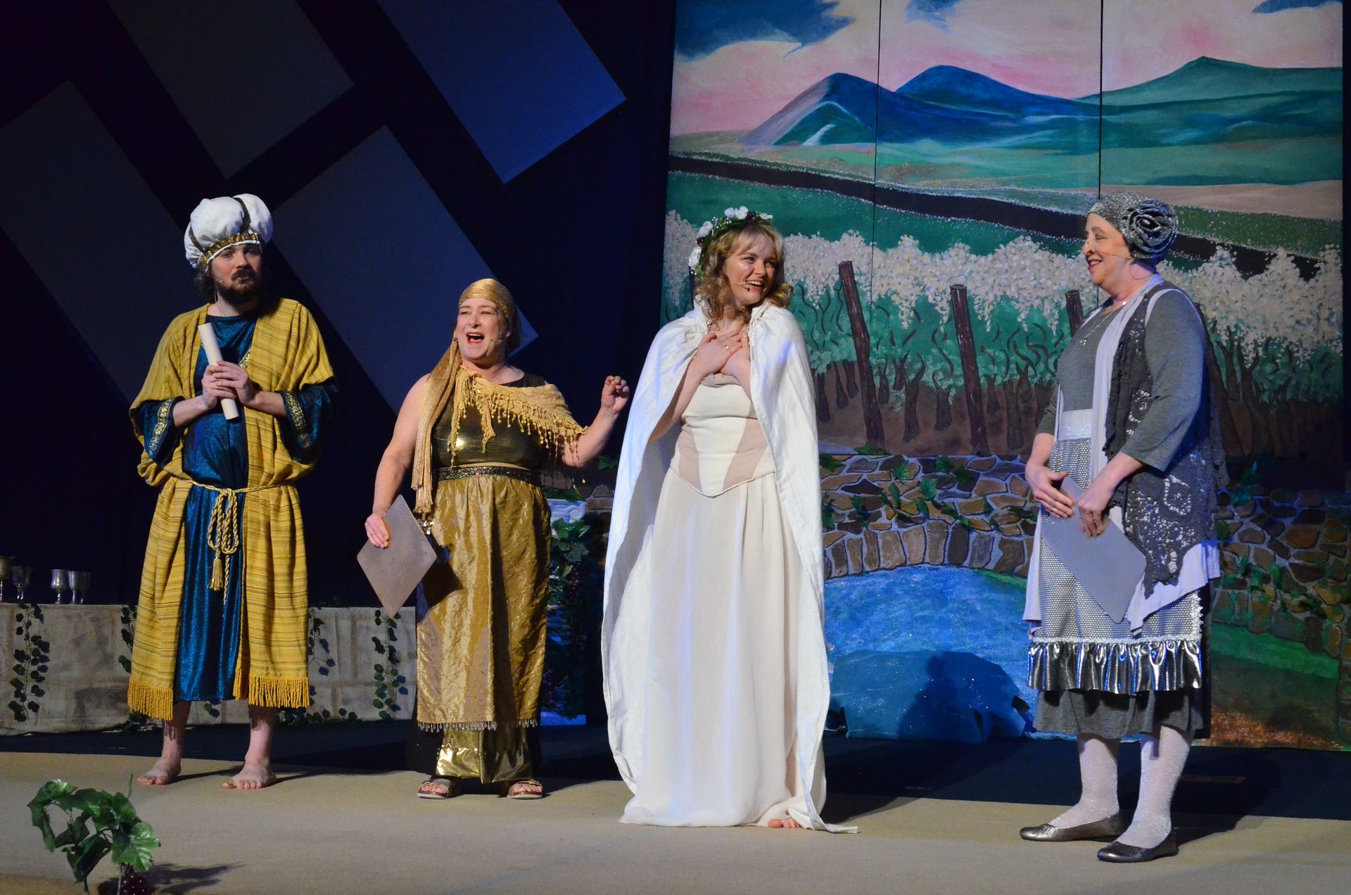 Four performers in colorful period costumes stand on stage against a painted backdrop depicting a mountainous landscape with fields and a blue body of water, with the performer in the center wearing a white draped gown and the others in gold and blue garments.