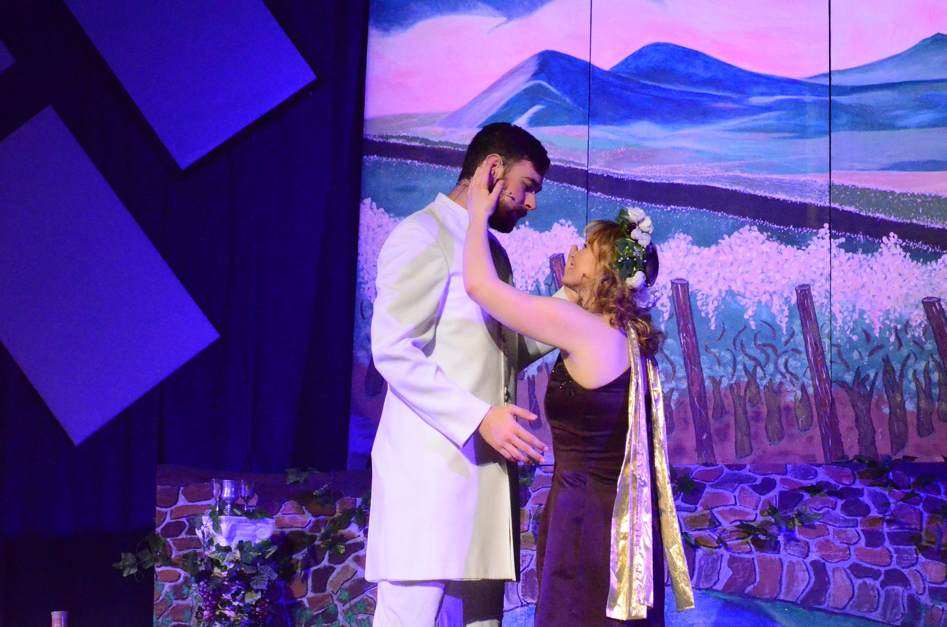 Two performers in formal attire dance together on stage against a vibrant backdrop of purple mountains and a pink sky, with dramatic blue lighting and stylized set pieces framing the scene.