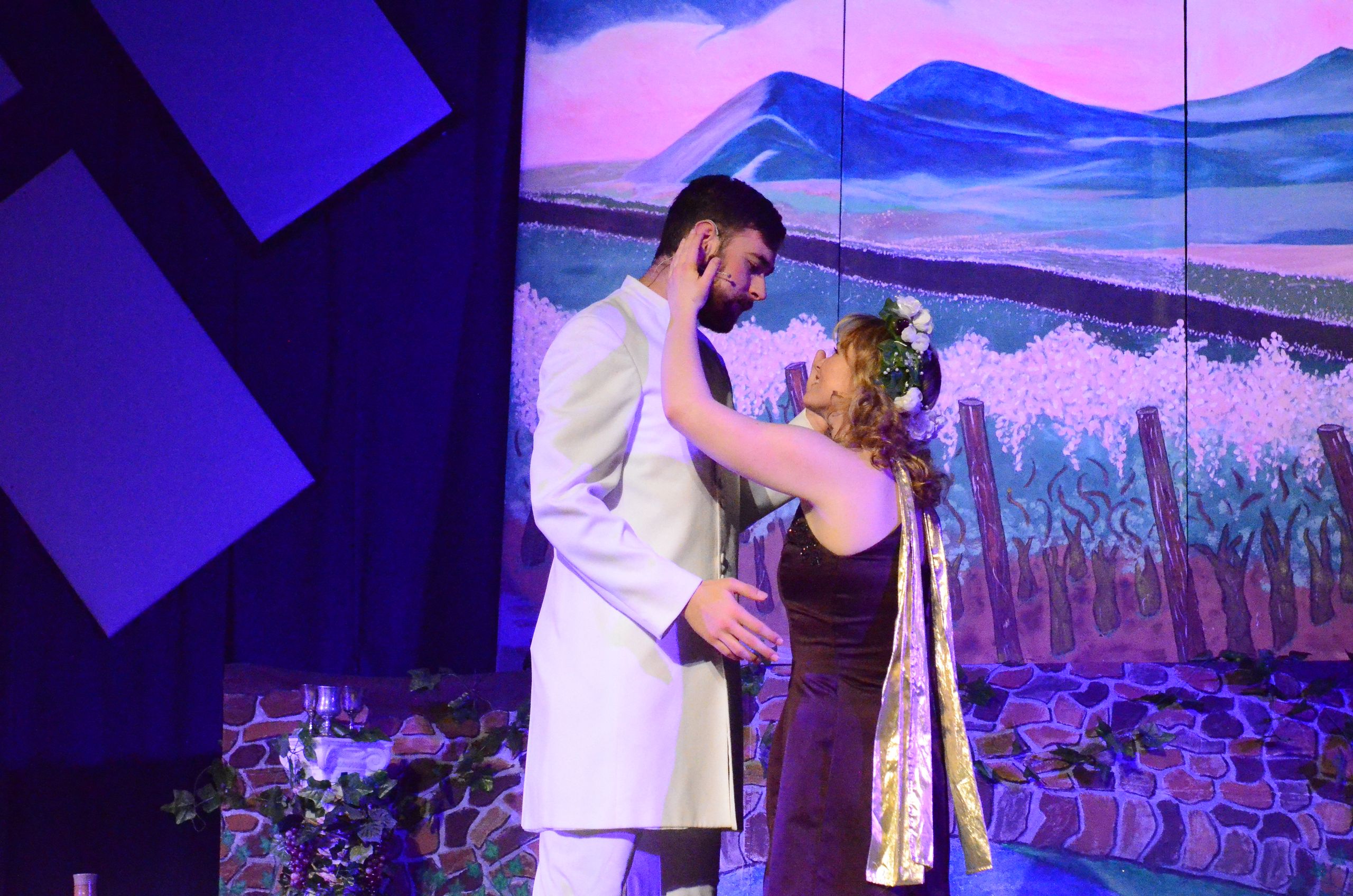 Two performers in formal attire dance together on stage against a vibrant backdrop of purple mountains and a pink sky, with dramatic blue lighting and stylized set pieces framing the scene.