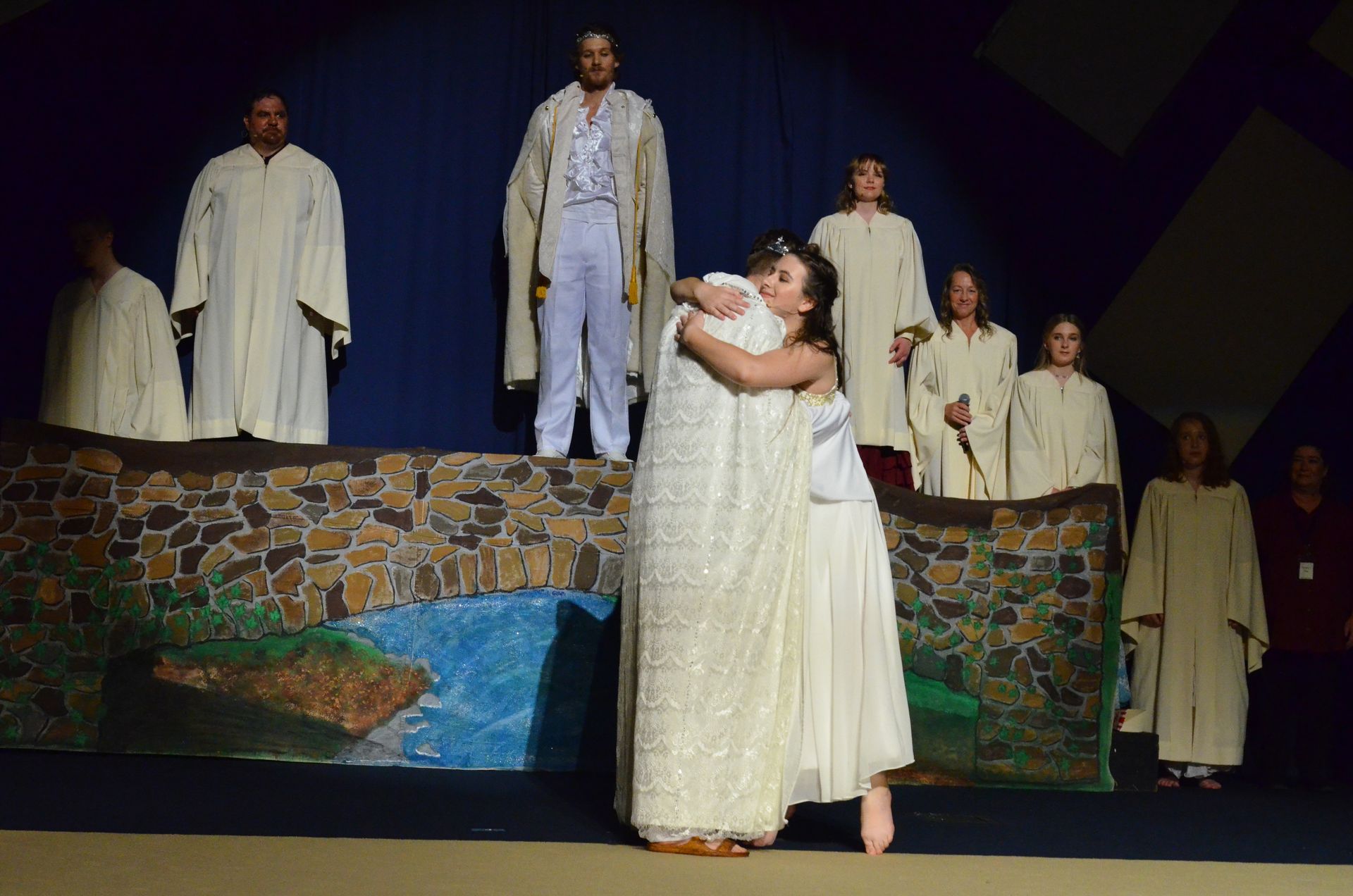 A cast of performers in cream-colored robes stands on stage around a painted backdrop of stone walls and water, while two performers in white garments embrace in the foreground.