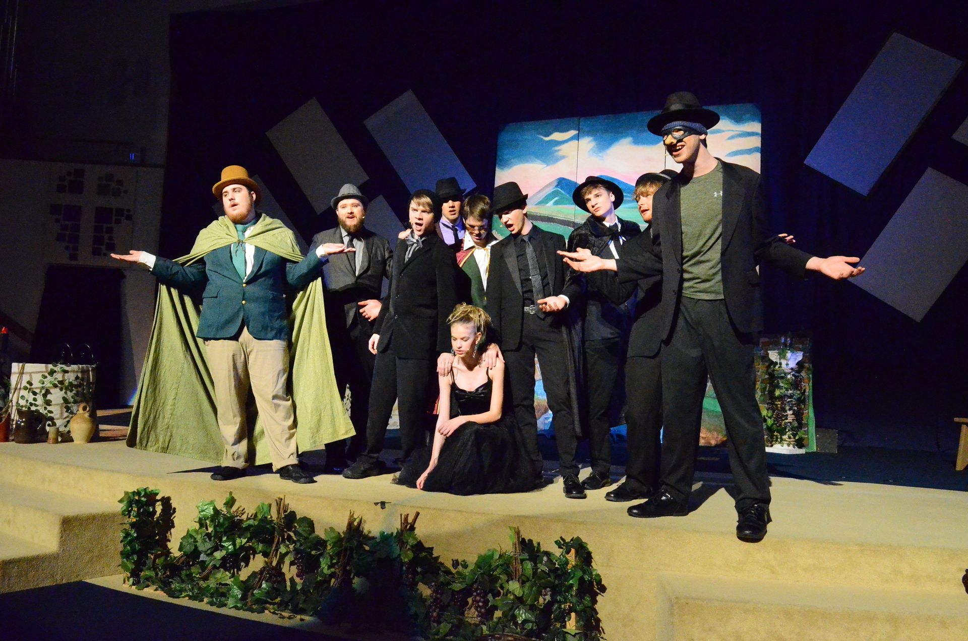 A cast of performers in period costumes including hats, capes, and suits stand together on stage with a painted backdrop, blue geometric set pieces, and potted plants, with one performer kneeling in the foreground.