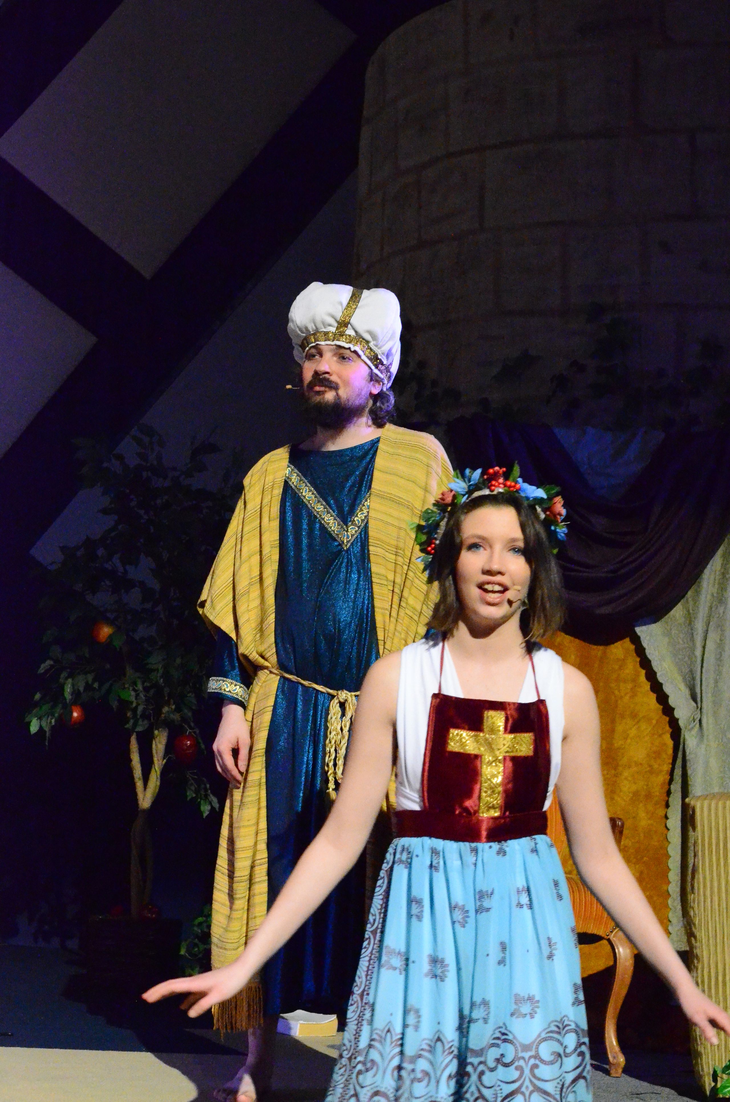 Two performers in colorful period costumes featuring yellow and blue fabrics with decorative gold trim stand on stage, one wearing a floral headdress and the other a white head covering, with a glowing golden cross visible on a burgundy panel behind them.