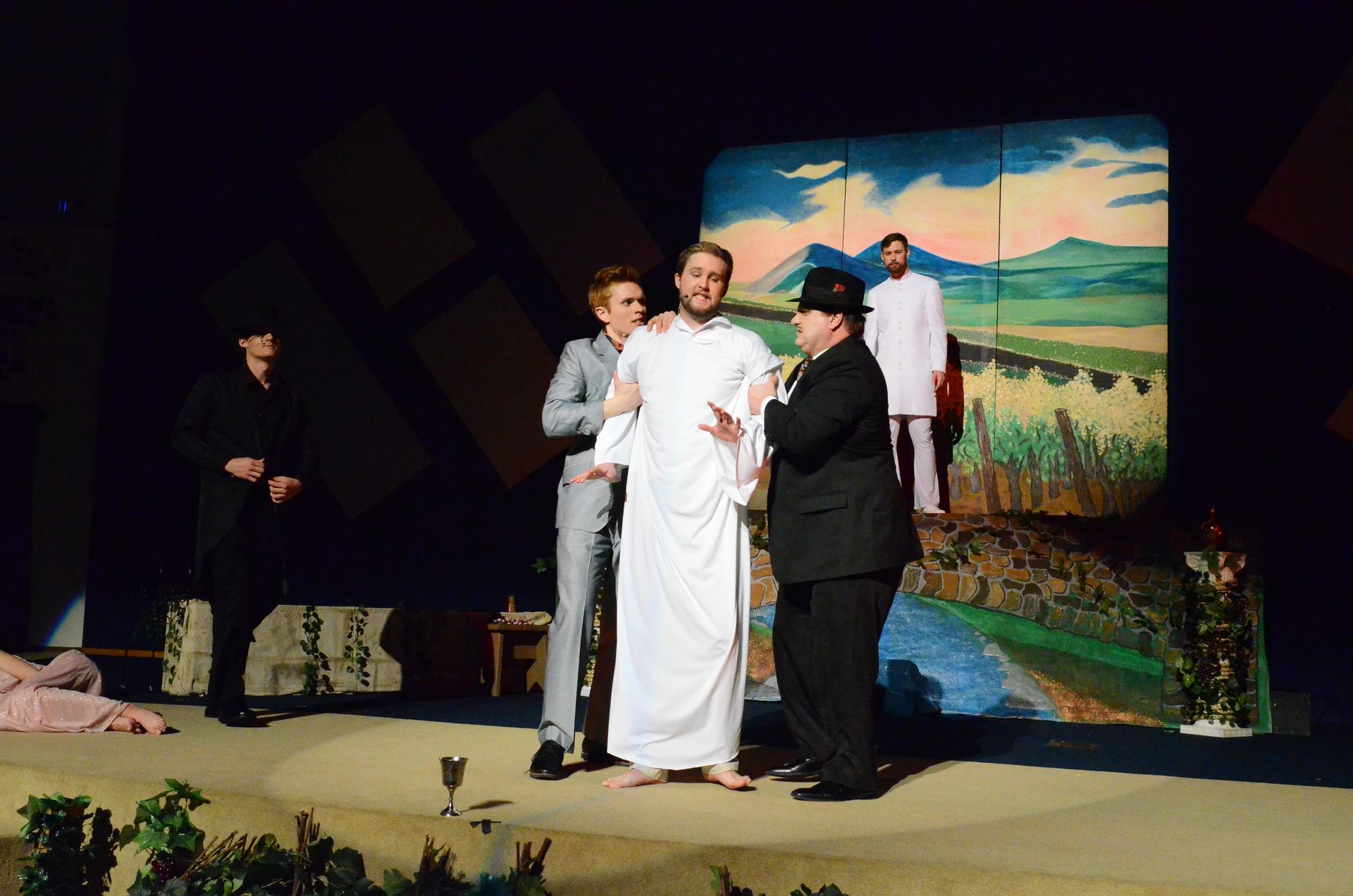 Multiple performers in period costumes interact on stage in front of a painted backdrop depicting a pastoral landscape with fields and sky, with additional cast members visible in the background.