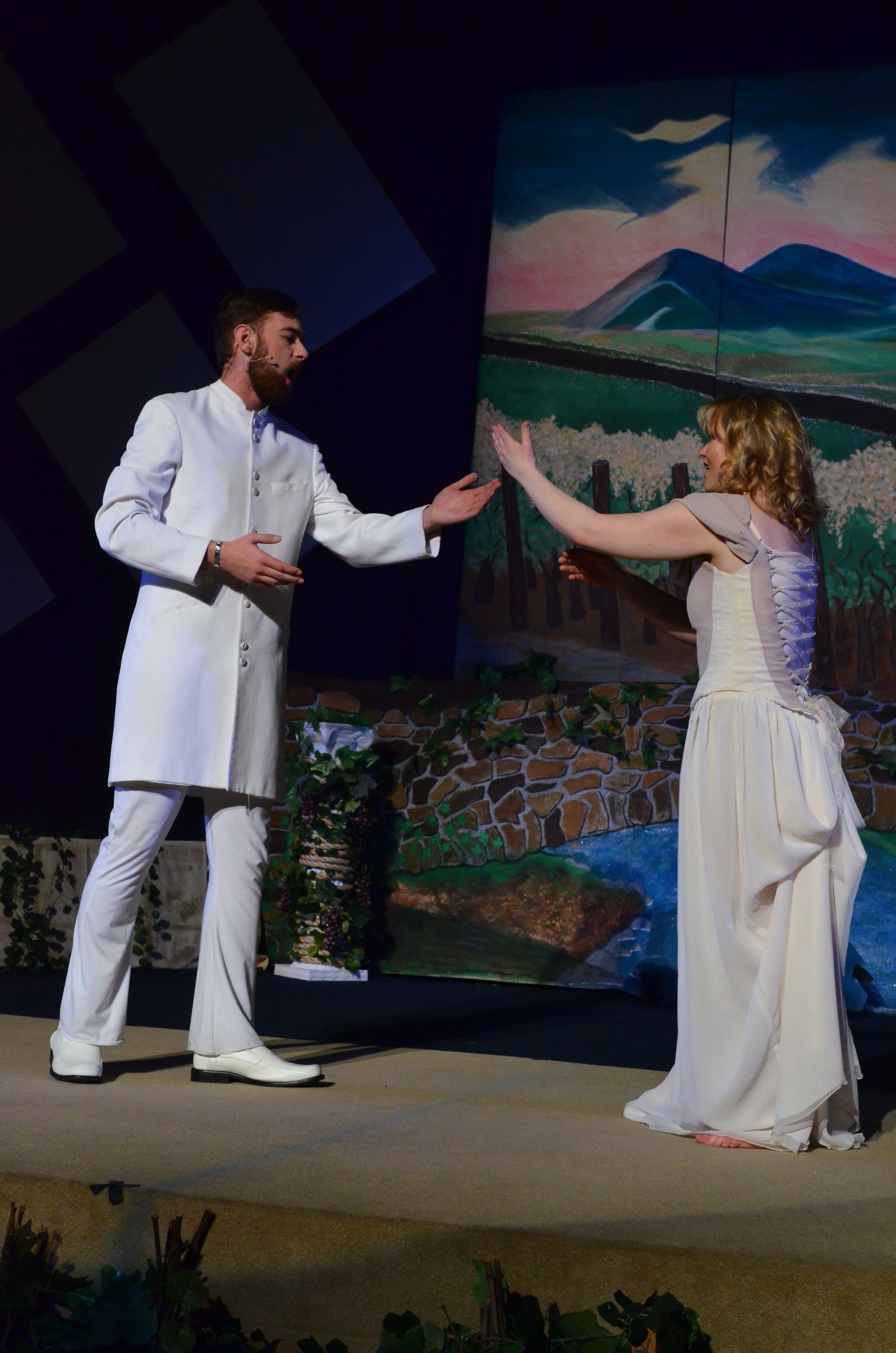 Two performers in white costumes stand on stage before a colorful backdrop depicting natural scenery, engaged in an emotional scene together.