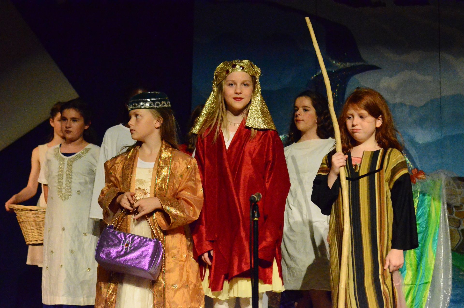 Five performers in elaborate biblical-era costumes stand on stage against a painted backdrop, with the center performer wearing a striking red robe and gold headdress while holding a microphone.