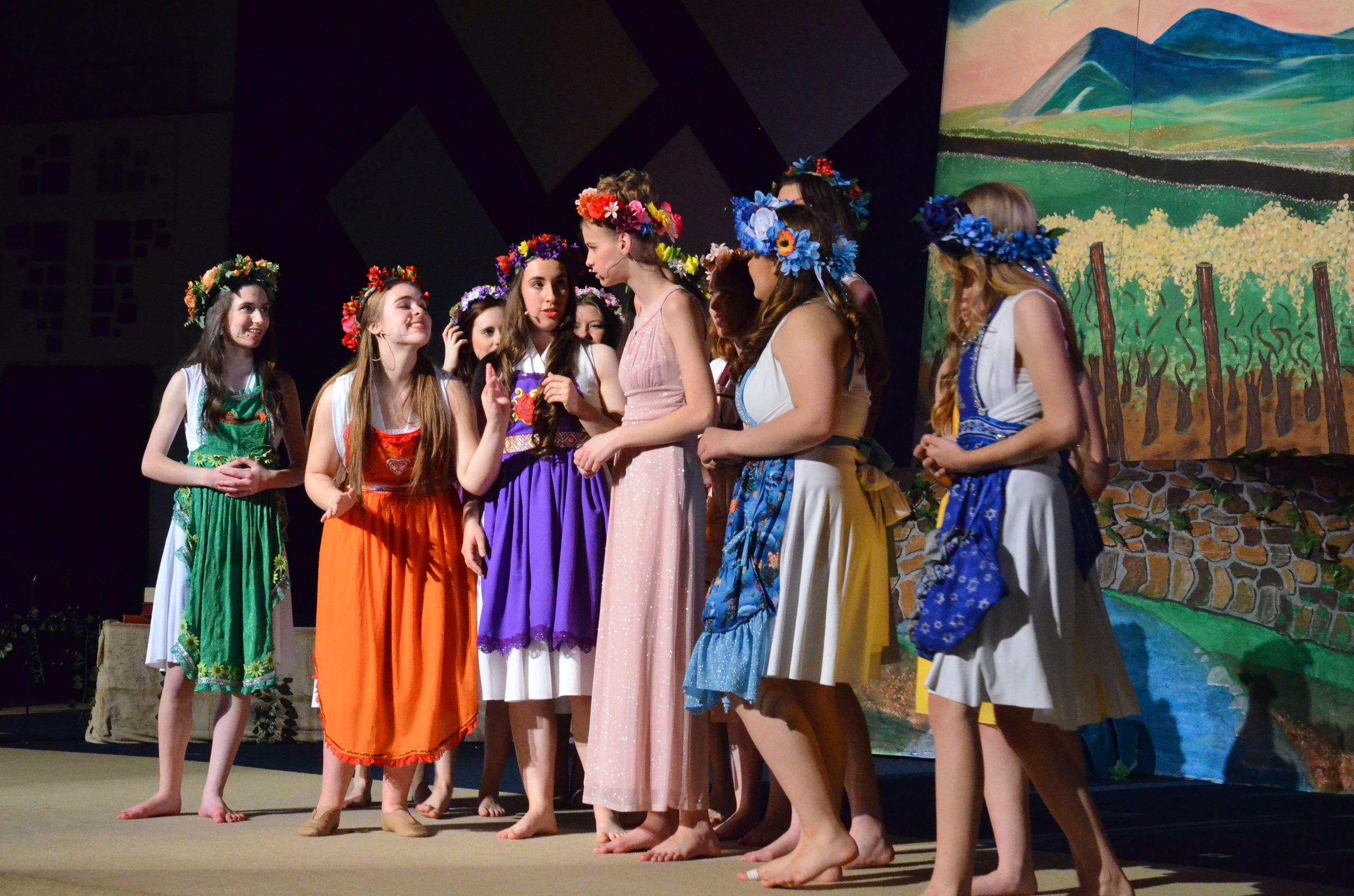 A cast of performers wearing colorful flower crowns and draped garments stands barefoot on stage in front of painted pastoral backdrops, engaging in an ensemble scene.