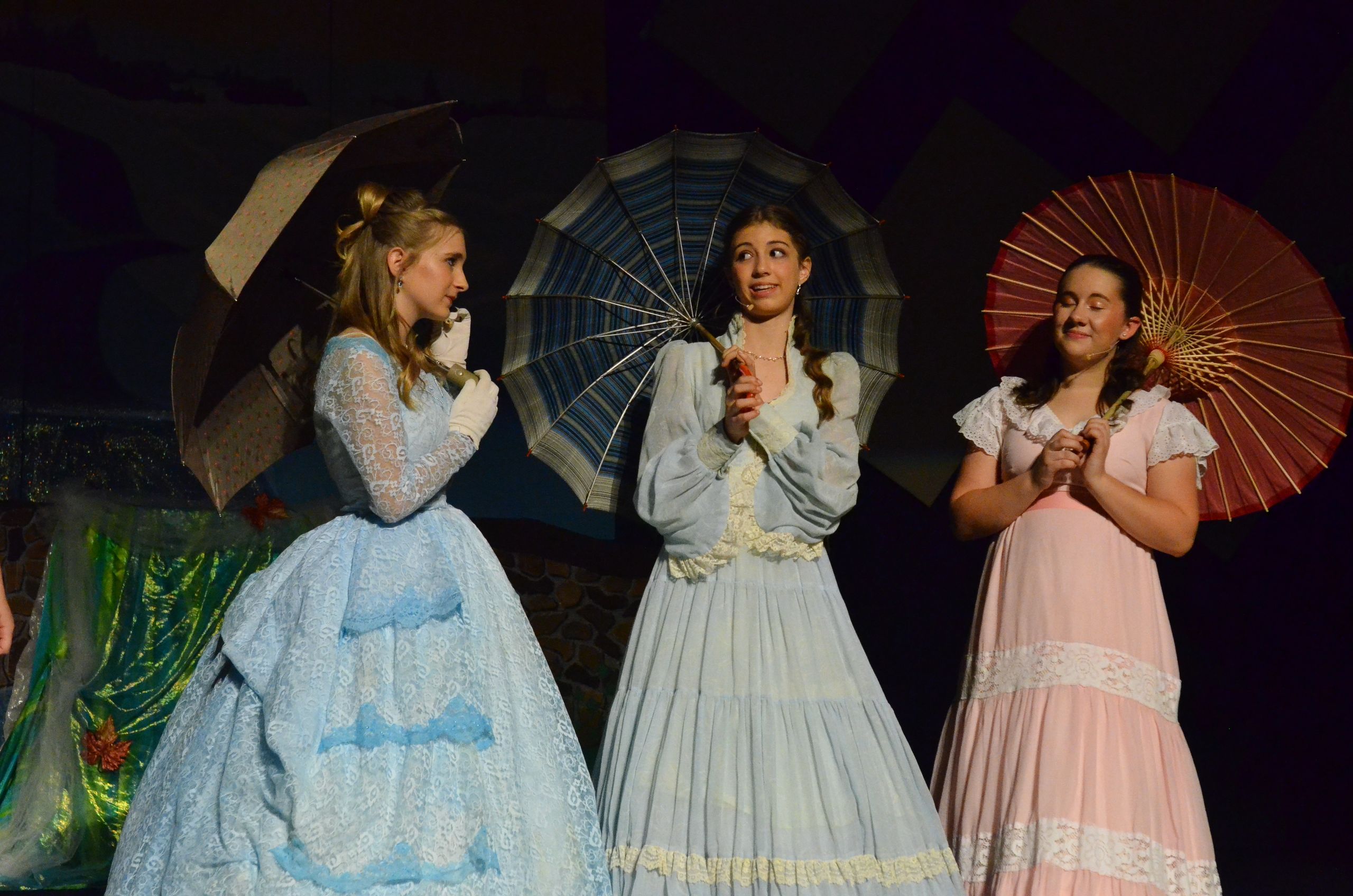 Three performers in period costumes hold decorative parasols while standing on stage against a dark background.