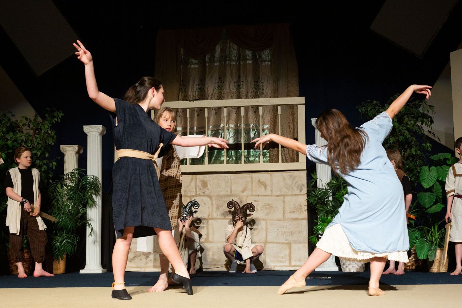 Performers in period costumes engage in an animated scene on a stage set with classical columns, potted plants, and a wooden door backdrop, with additional cast members positioned around the edges of the set.