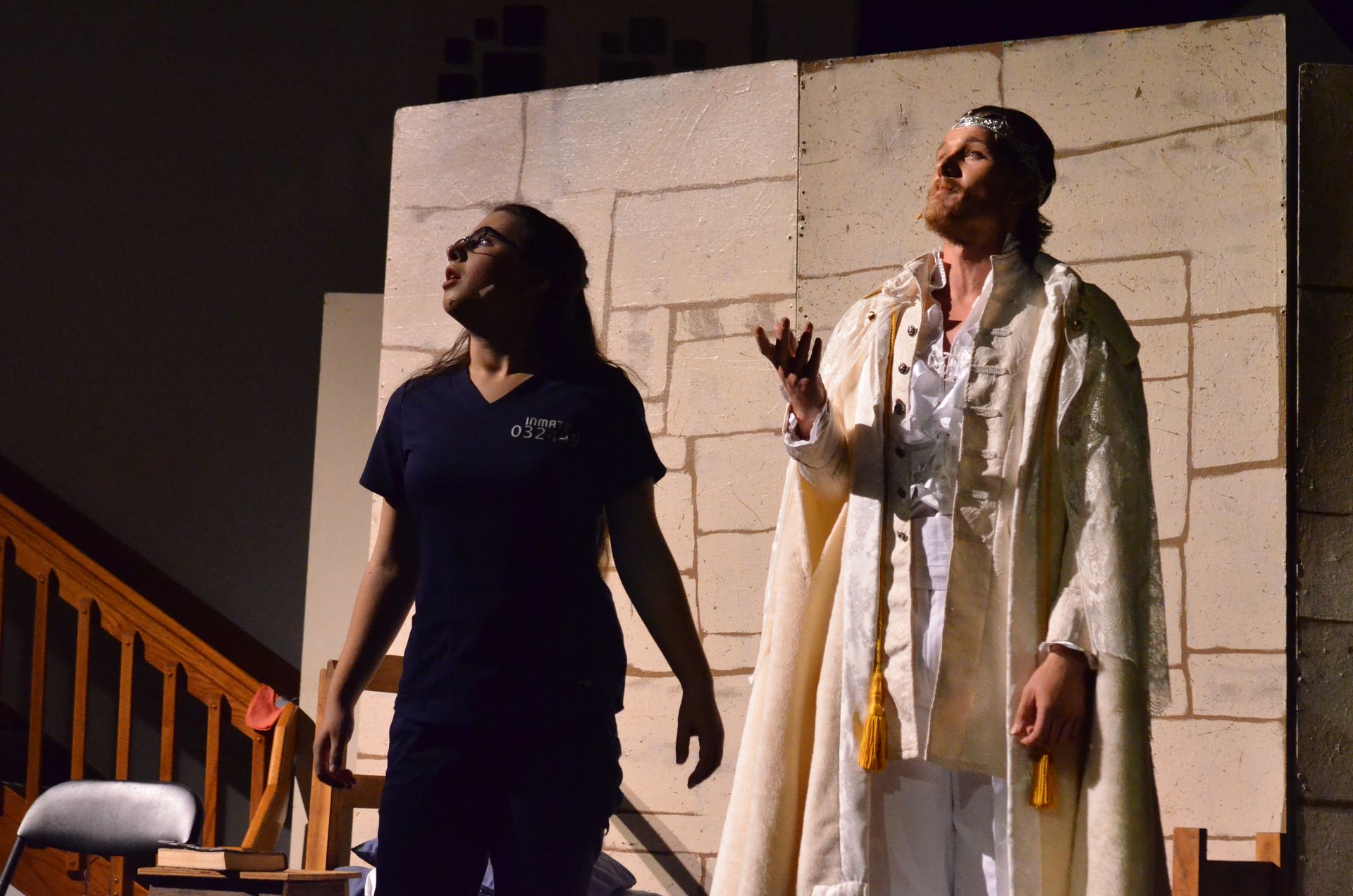 Two performers stand on stage against a stone wall backdrop, with one wearing a dark t-shirt and the other wearing a long cream-colored robe with gold accents and a draped outer garment, gesturing expressively during the scene.