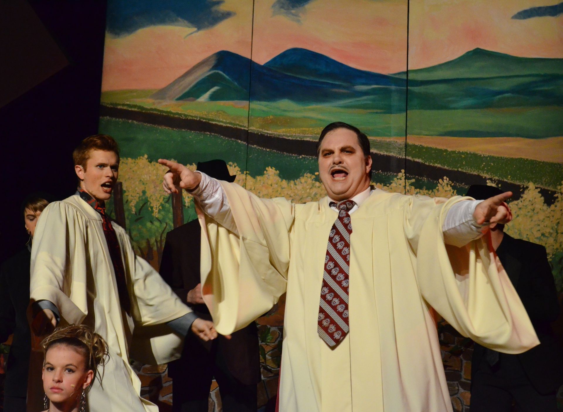 A performer in a cream-colored robe and patterned tie gestures expressively with arms outstretched while singing, surrounded by other cast members in period costumes against a painted backdrop of rolling hills and mountains.