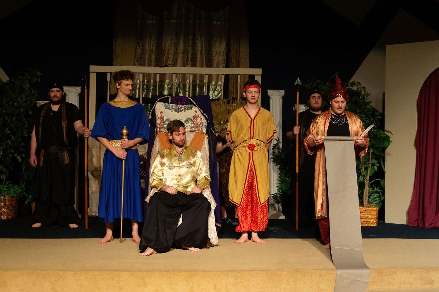 A cast of performers in elaborate biblical-era costumes stand on a stage set with classical columns, potted plants, and draped backdrops, with one performer seated centrally wearing an ornate gold and white robe.