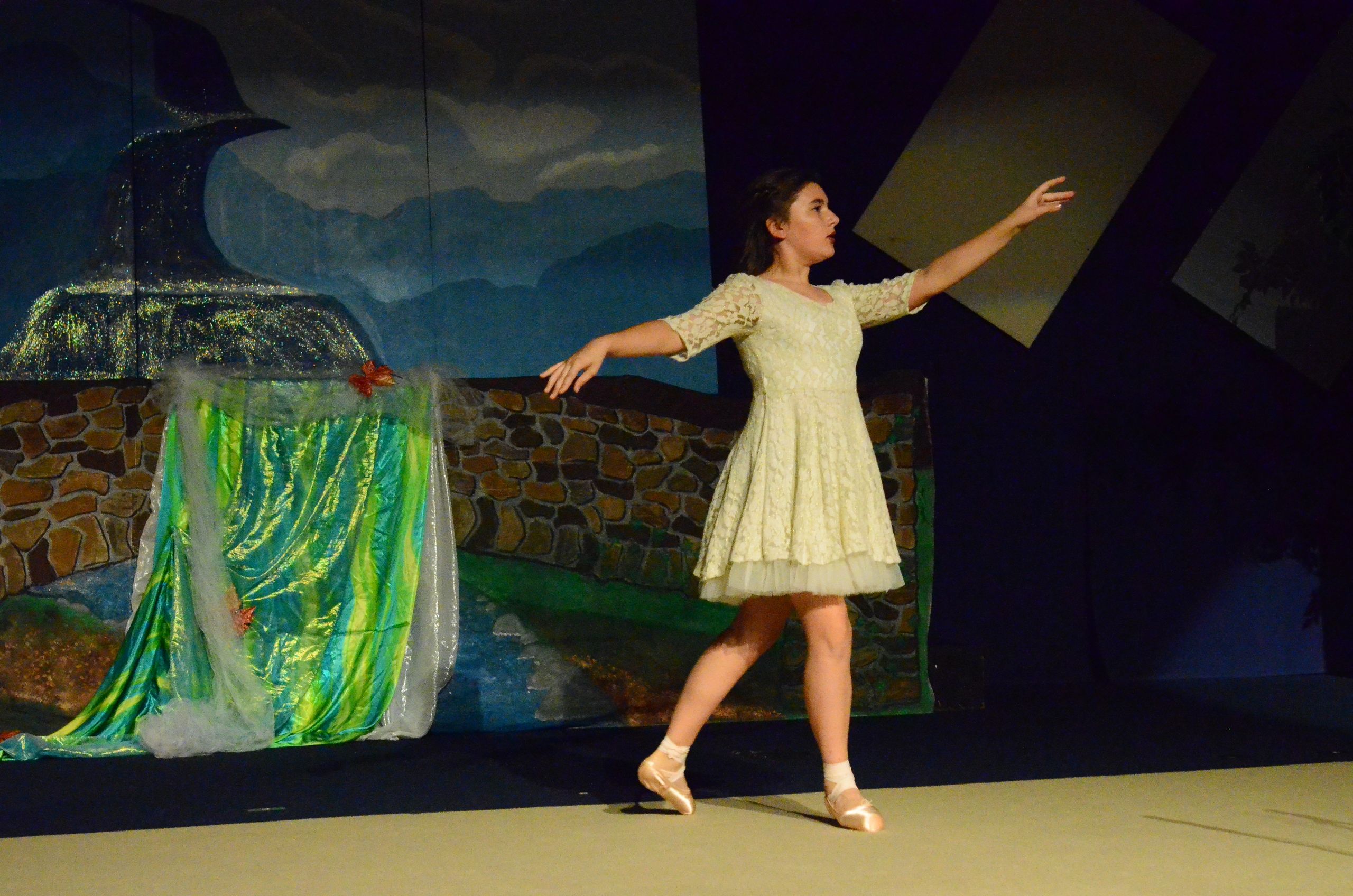 A performer in a cream-colored dress stands on stage with one arm extended, positioned beside an ornate prop decorated with green and gold fabric, with a painted backdrop depicting a moonlit mountainous landscape behind them.
