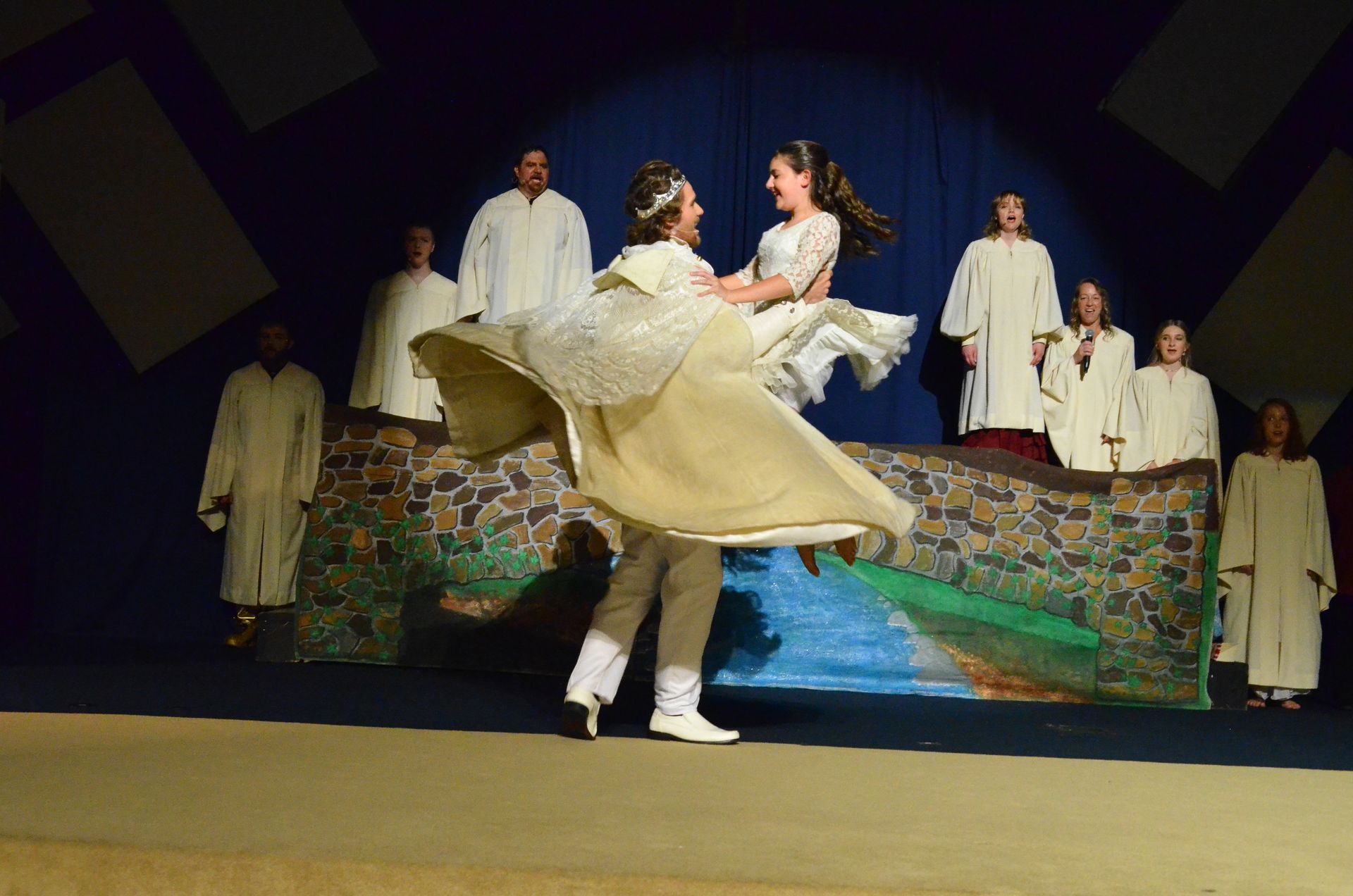 A performer in an elegant cream-colored gown with a flowing cape dances with another performer while surrounded by cast members dressed in simple robes on a colorful stone-patterned set against a deep blue backdrop.