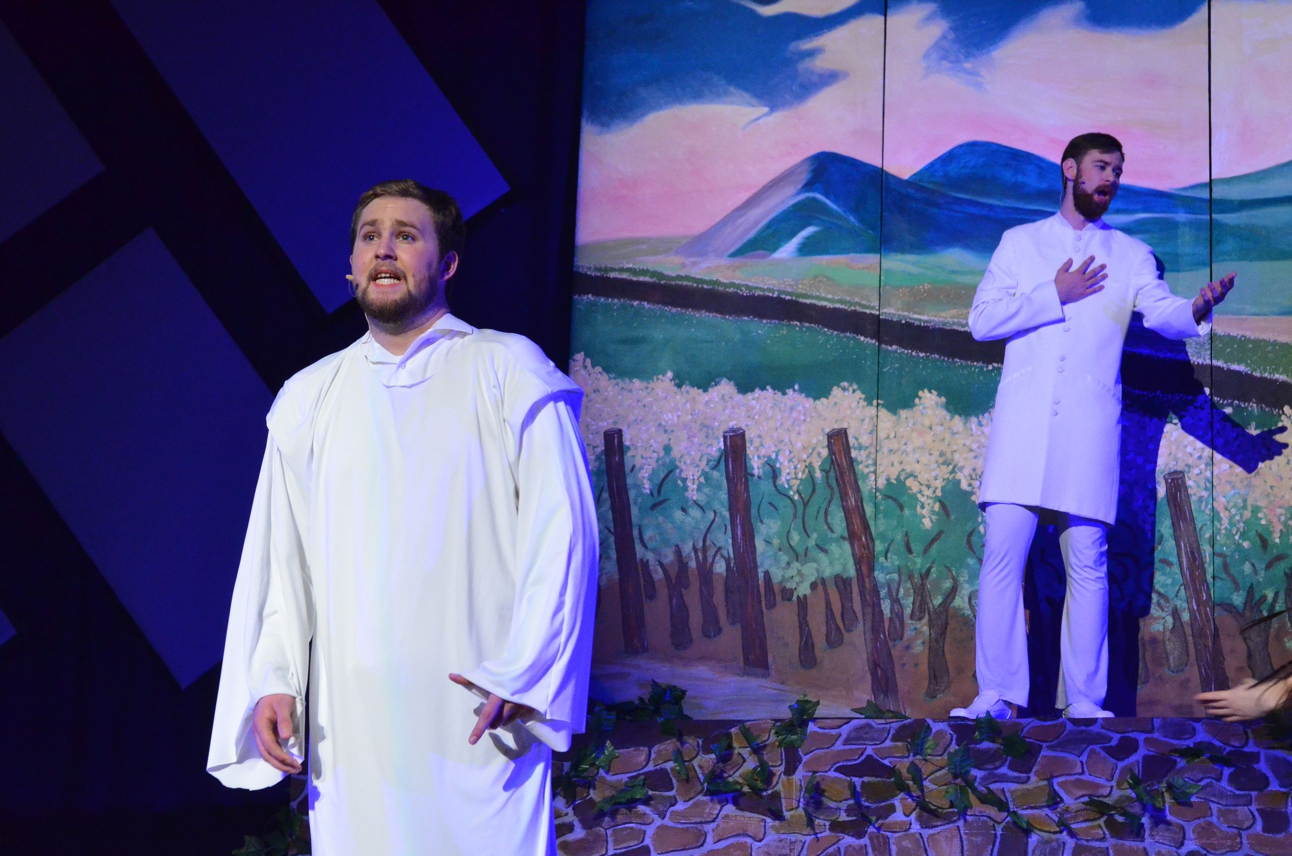 Two performers in white robes stand on stage against a painted backdrop depicting a biblical landscape with mountains, fields, and wooden posts, with blue-tinted theatrical lighting illuminating the scene.