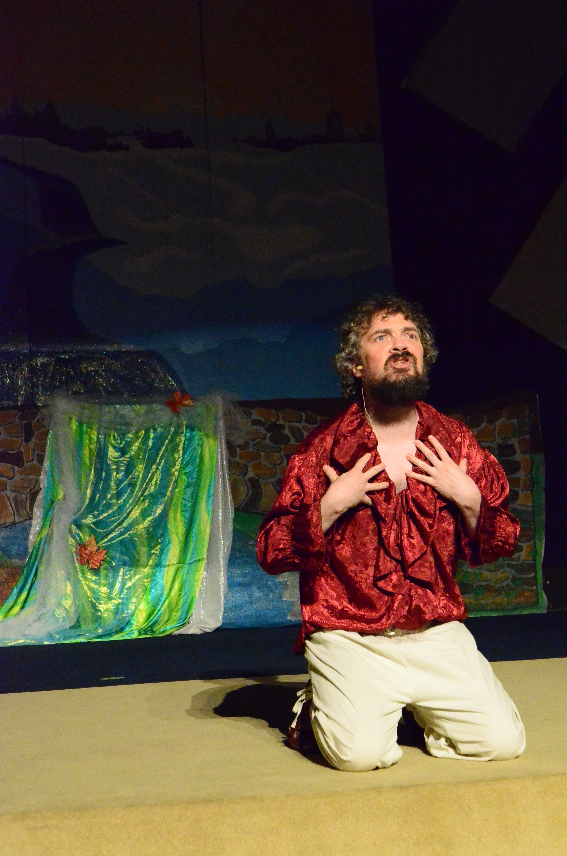 A performer in a red sequined costume lies on a colorful painted stage set while another performer in cream-colored clothing reclines above, with dark drapes and yellow walls visible in the theatrical setting.