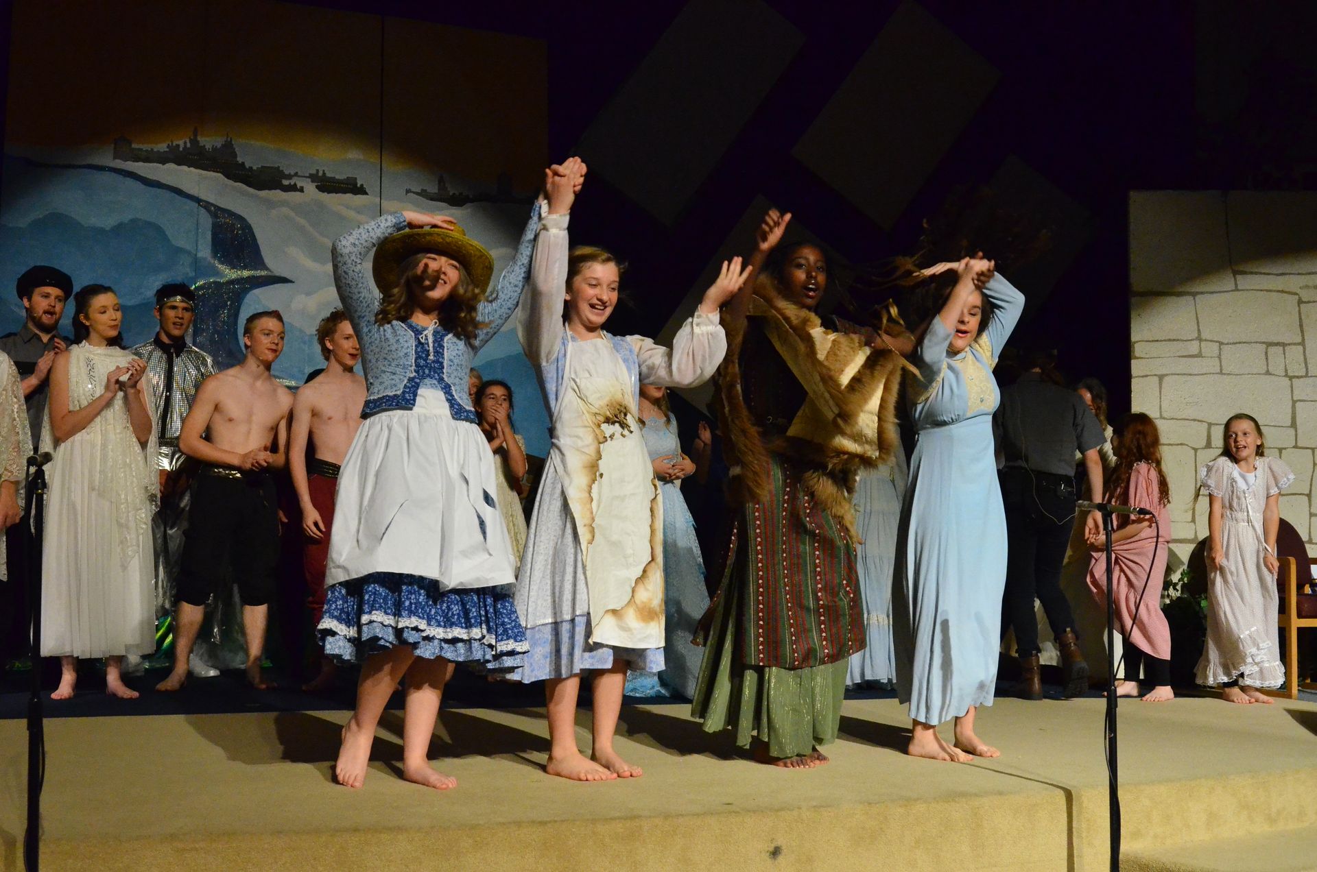 A diverse ensemble of performers in period costumes stands barefoot on stage with raised arms in celebration, with a painted backdrop depicting a coastal biblical scene behind them.