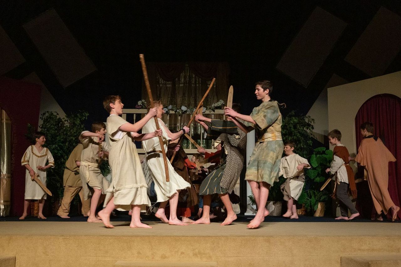 The cast performs a spirited scene with wooden staffs and props on stage, wearing earth-toned costumes and bare feet, surrounded by painted set pieces and green foliage.