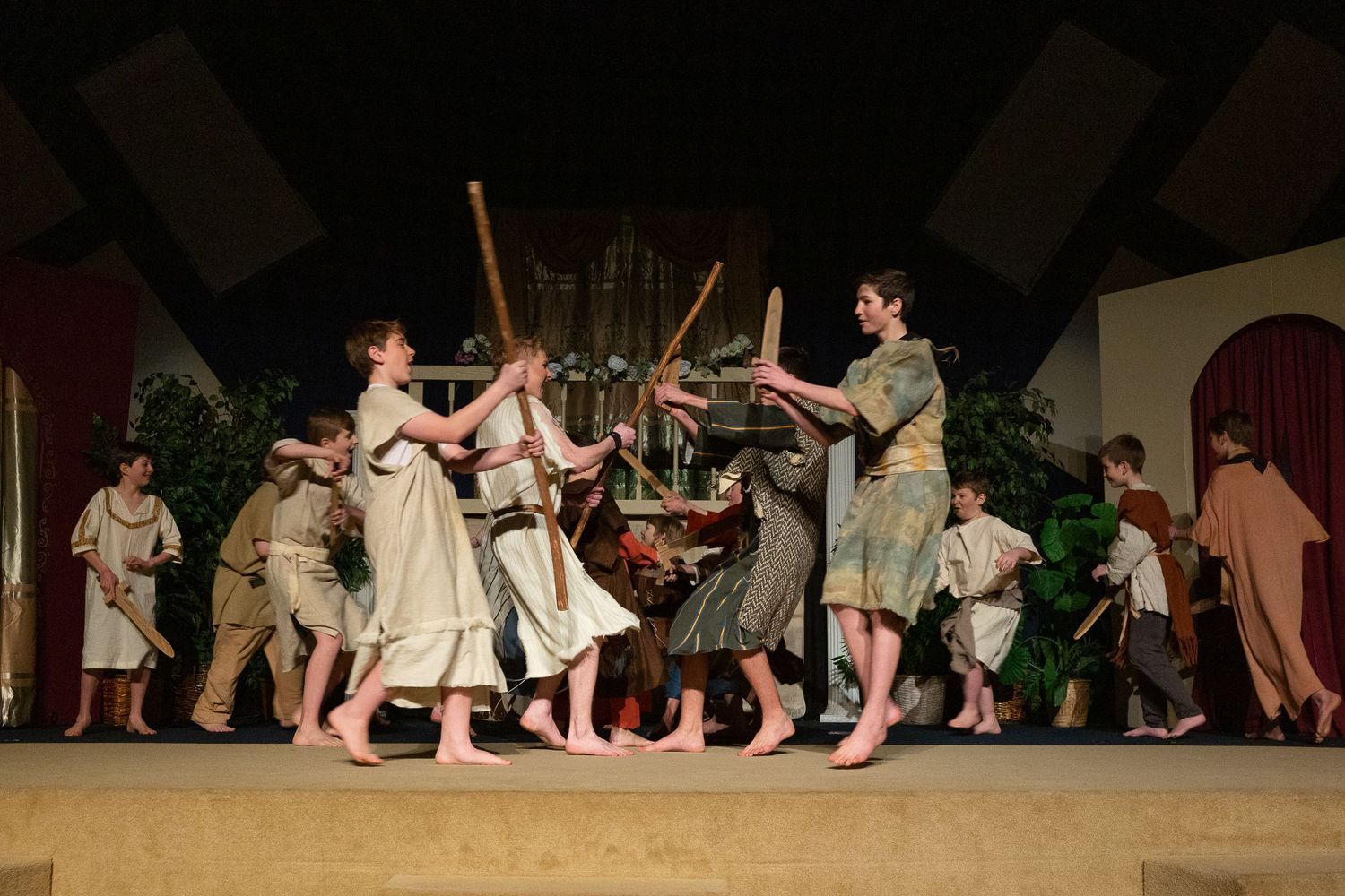 The cast performs a spirited scene with wooden staffs and props on stage, wearing earth-toned costumes and bare feet, surrounded by painted set pieces and green foliage.