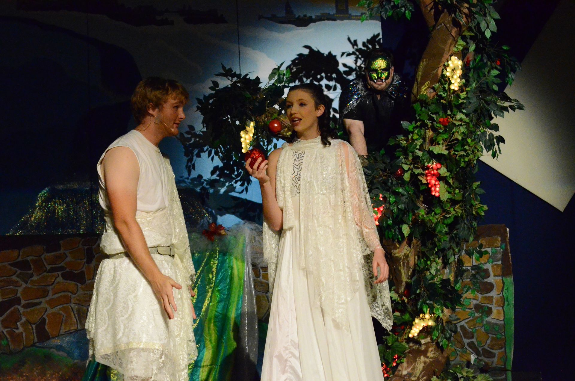 Two performers in white draped costumes stand in a garden setting decorated with flowering vines and greenery, with one holding a red apple while a figure in elaborate costume stands behind them.