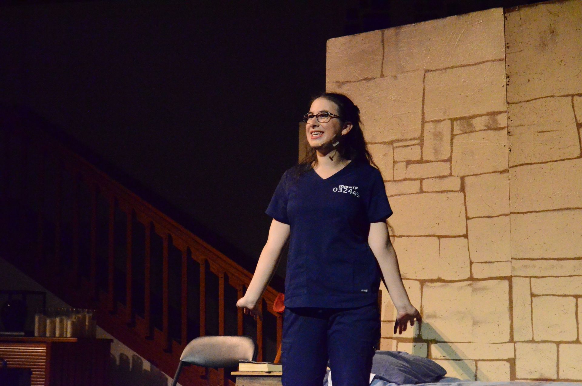 A performer wearing glasses and a dark blue shirt stands on a stage with tan brick walls and wooden railings, smiling while gesturing expressively during what appears to be a theatrical performance.
