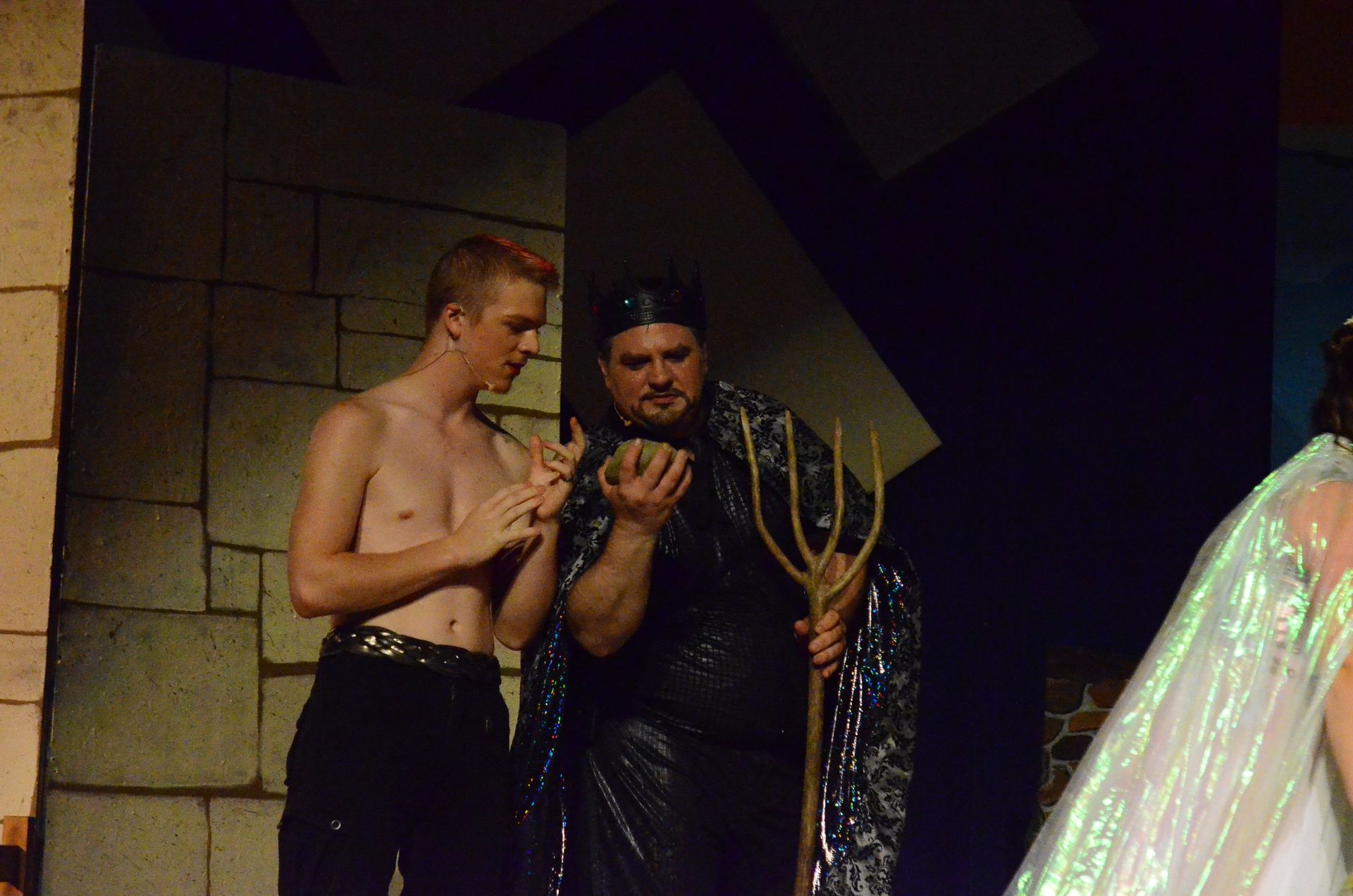 Two performers in elaborate costume stand together on stage against a stone wall backdrop, with one wearing a black sequined robe and holding a golden staff while the other stands shirtless beside them.