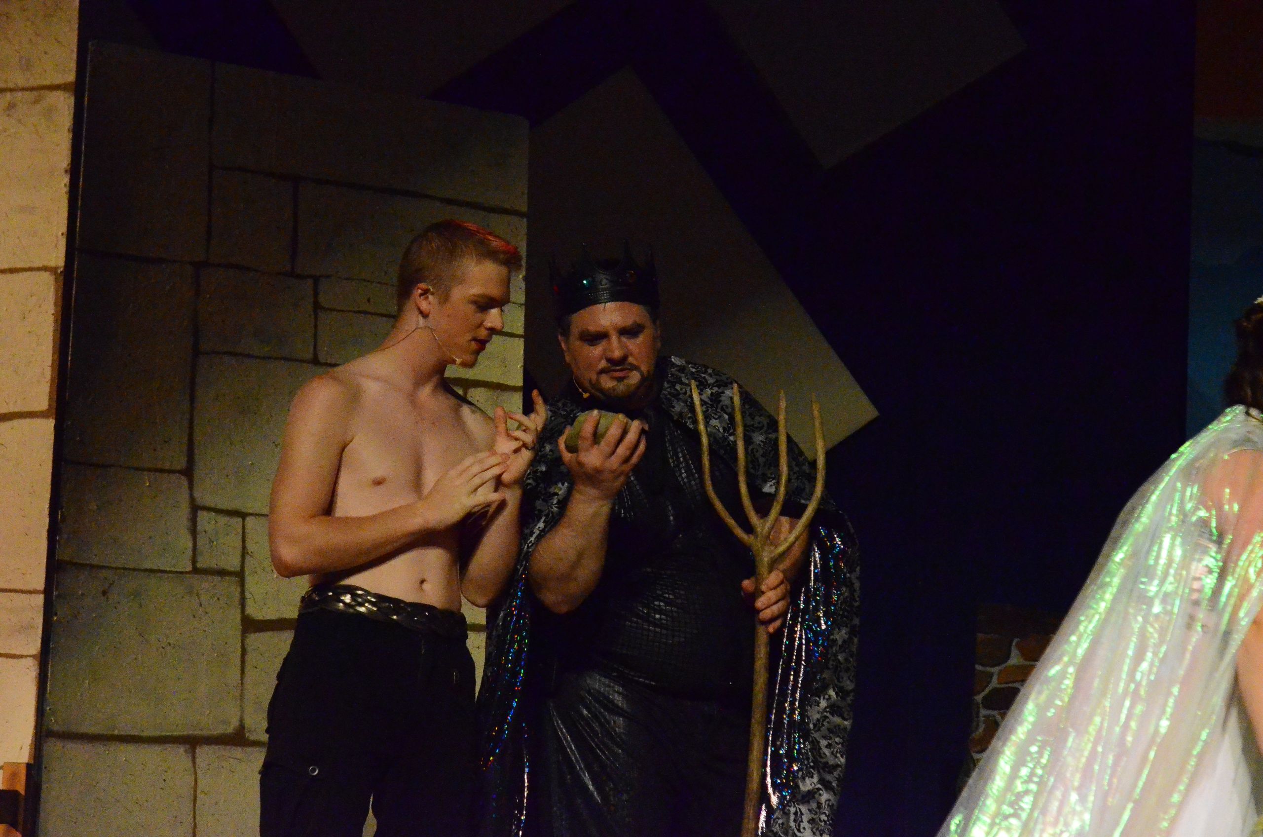 Two performers in elaborate costume stand together on stage against a stone wall backdrop, with one wearing a black sequined robe and holding a golden staff while the other stands shirtless beside them.