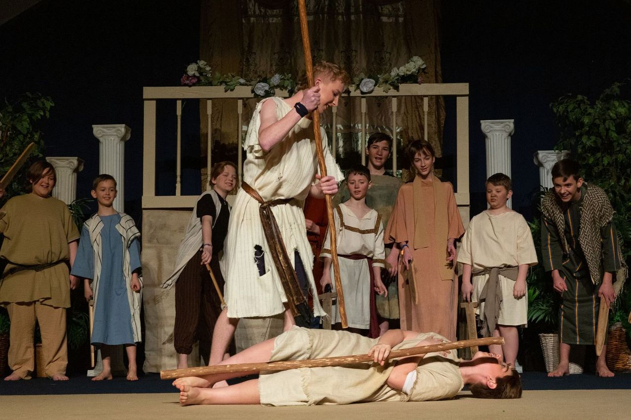 A cast of performers in biblical-era costumes stands on a wooden stage set with classical columns, while one performer wielding a staff stands elevated above another performer lying prone on the ground.