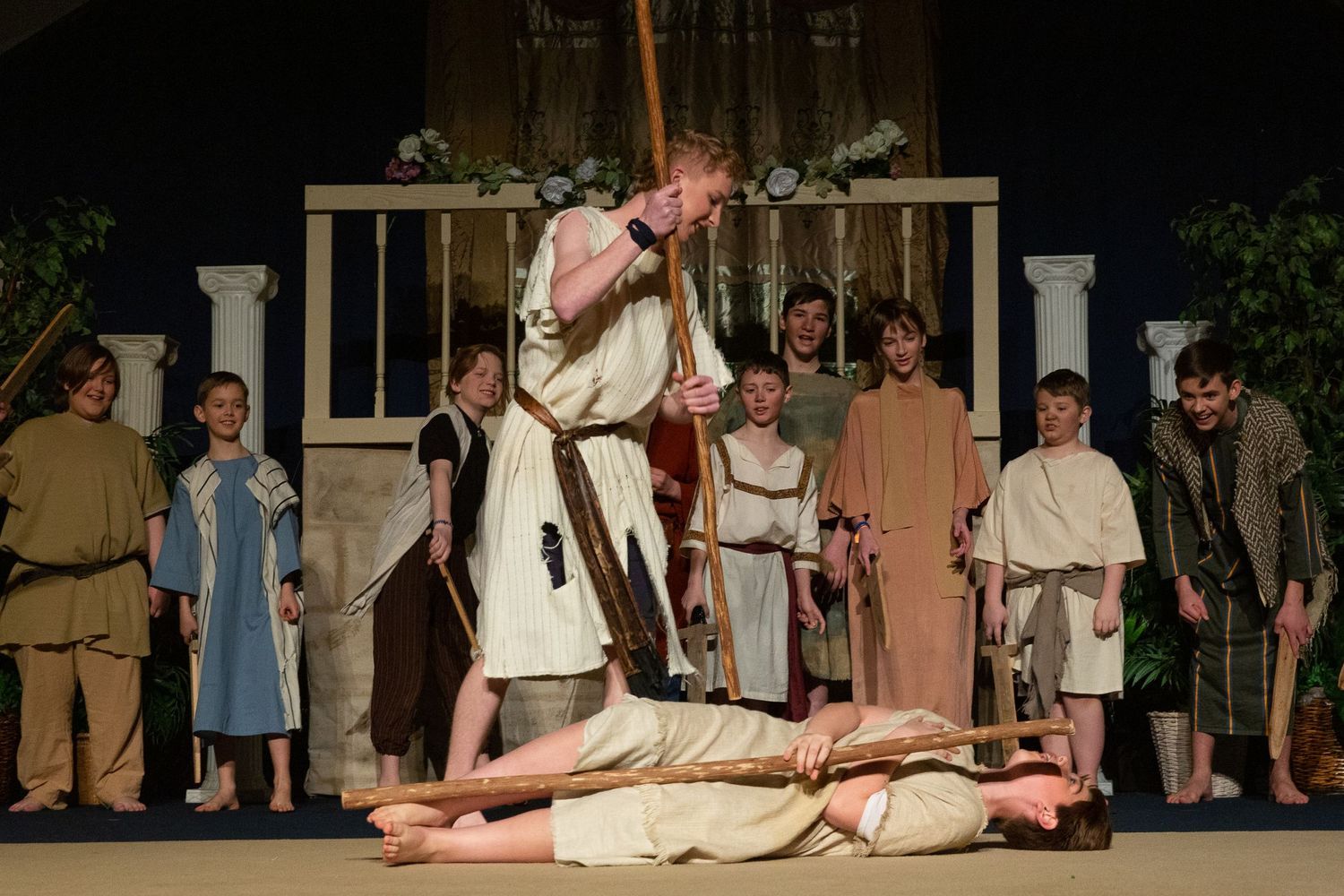 A cast of performers in biblical-era costumes stands on a wooden stage set with classical columns, while one performer wielding a staff stands elevated above another performer lying prone on the ground.