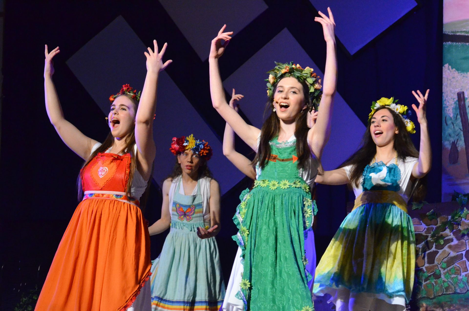 Five performers wearing colorful floral-adorned dresses stand on stage with raised arms and joyful expressions against a blue geometric backdrop.
