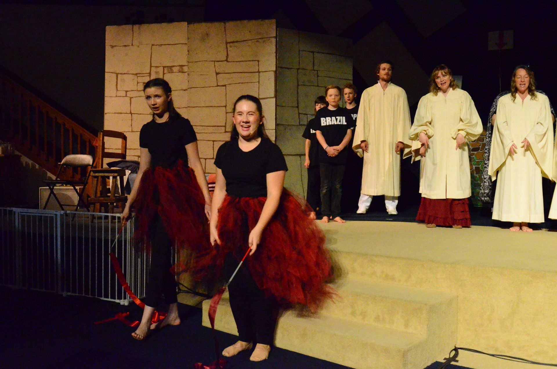 Two performers in black shirts and voluminous red skirts dance on stage while cast members in cream-colored robes stand behind them, with a stone wall backdrop and wooden furniture visible to the left.