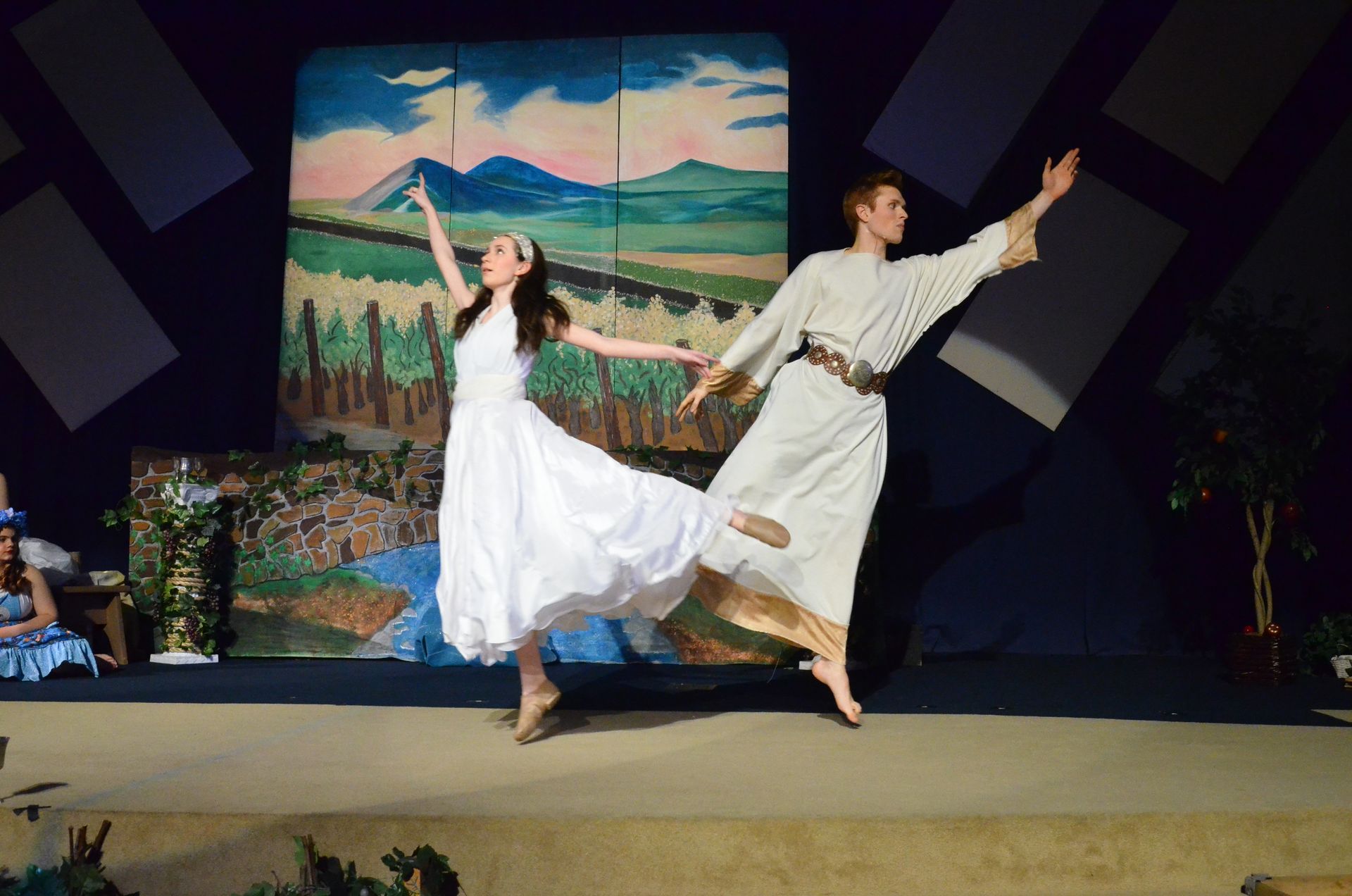 Two performers in white flowing garments dance expressively on stage in front of a painted backdrop depicting mountains and farmland, with their arms extended in joyful movement.
