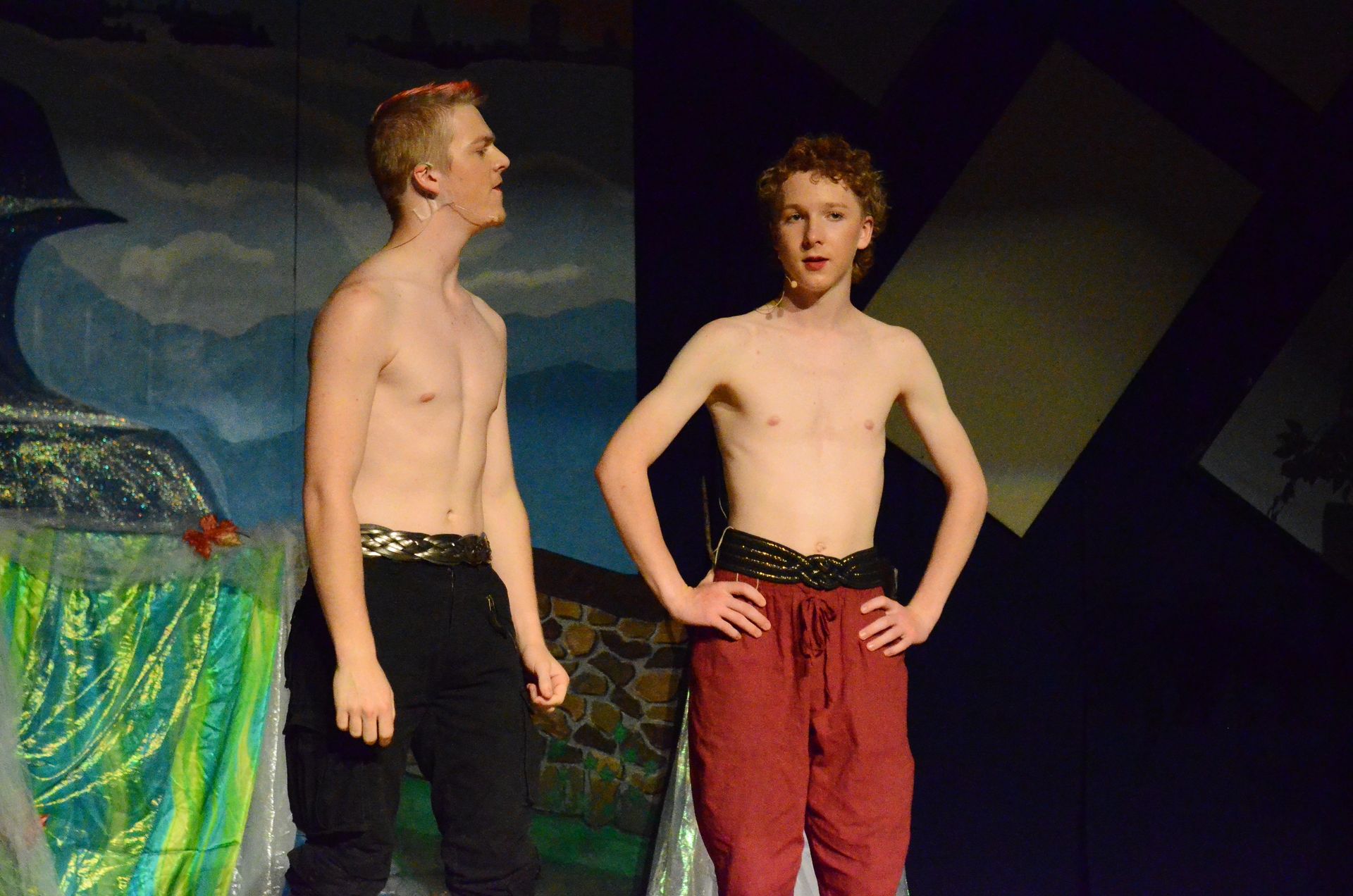 Two shirtless performers stand on stage against a colorful backdrop featuring blue sky and green foliage, wearing decorative waistbands and colored pants in black and burgundy.