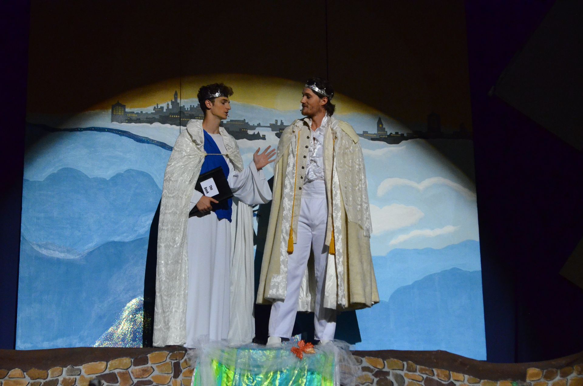 Two performers in elaborate period costumes with flowing cloaks stand on stage conversing against a backdrop depicting mountains and a distant cityscape.