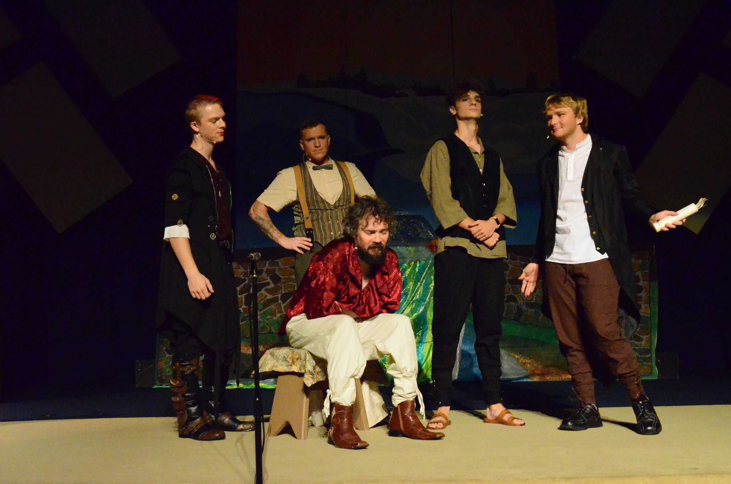Five performers stand on a stage in period costumes—including a red silk garment, a striped vest, and earth-toned clothing—with one seated while others stand around him, positioned in front of a painted backdrop depicting a Middle Eastern landscape.